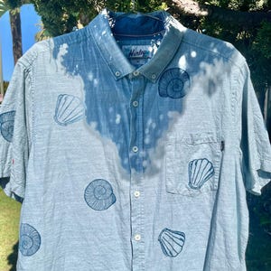 Puede incluir: Camisa azul claro de manga corta con estampado de conchas marinas. La camisa presenta un diseño decolorado y el logotipo de Hurley en el cuello. Tiene un bolsillo en el pecho y está colgada de una percha metálica.