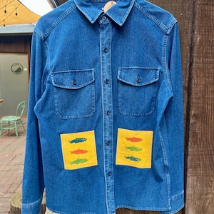 May include: A blue denim long-sleeve button-down shirt featuring a classic collar and two chest pockets. The front lower section includes two rectangular yellow patches, each decorated with three small, colorful fish illustrations in red, green, and orange.
