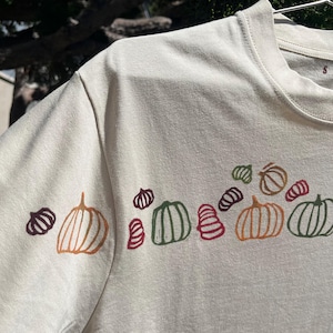 以下が含まれることがあります： オレンジ、緑、バーガンディの色合いで、カラフルなカボチャのイラストが並んだクリーム色のTシャツ。カボチャは様式化され、胸に配置されています。Tシャツは丸いネックラインと半袖です。