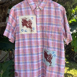 Pode incluir: Uma camisa de manga curta com um padrão xadrez em rosa, amarelo e azul. Possui dois patches com uma ilustração de cavalo marrom e ferraduras azuis. A camisa está pendurada em um cabide de madeira.