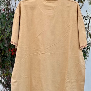 May include: A tan t-shirt with a red and brown embroidered pattern around the neckline. The pattern is a repeating design of a stylized "U" shape.
