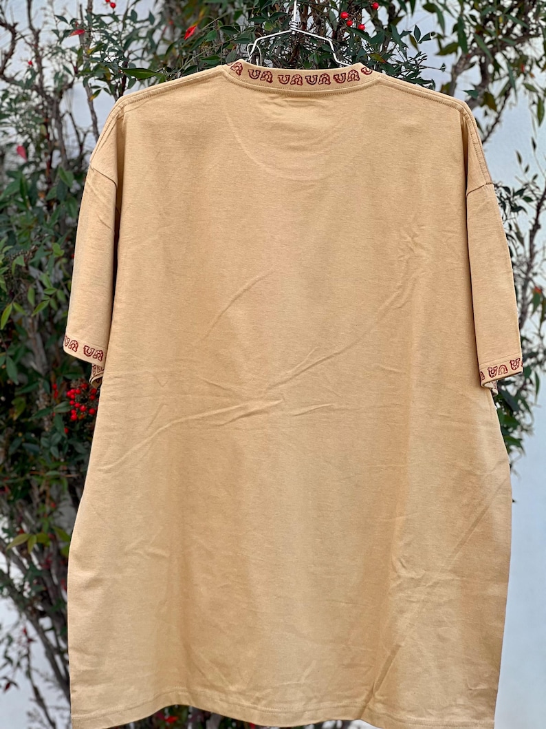 May include: A tan t-shirt with a red and brown embroidered pattern around the neckline. The pattern is a repeating design of a stylized "U" shape.