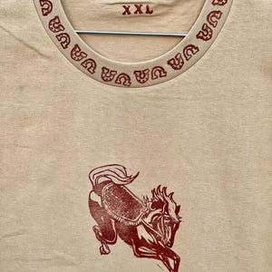May include: A tan t-shirt with a red horse design and a red horseshoe pattern around the neckline. The shirt has a size tag that says "XXL".