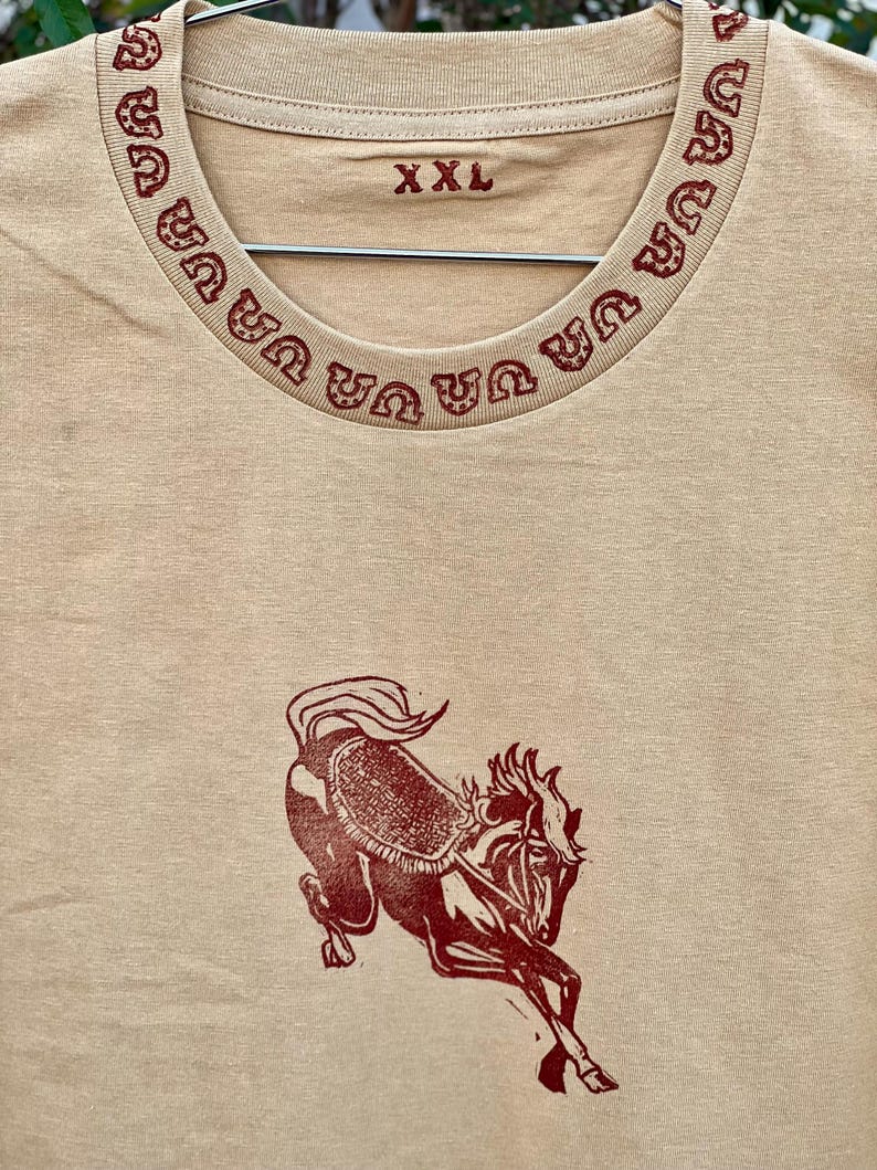 May include: A tan t-shirt with a red horse design and a red horseshoe pattern around the neckline. The shirt has a size tag that says "XXL".