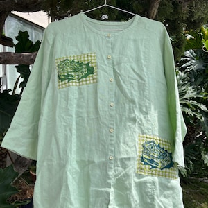 Peut inclure: Une chemise boutonnée vert clair à manches trois-quarts et encolure ronde. La chemise présente deux poches plaquées carrées, une sur la poitrine droite et une en bas à gauche, chacune avec une bordure à carreaux vichy verts et blancs.