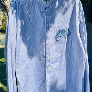 Puede incluir: Camisa abotonada azul claro con rayas blancas verticales. Presenta un pequeño diseño bordado de langosta en el bolsillo y una "J" en cursiva en el lado izquierdo. La camisa está colgada en una percha.