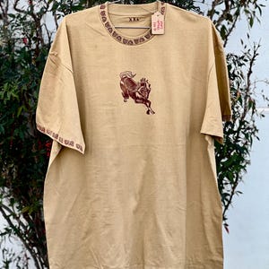 May include: A tan t-shirt with a brown horse graphic and a red trim around the neck and sleeves. The shirt has a tag that says "@Get True Cozy" and "XXL" printed on it.