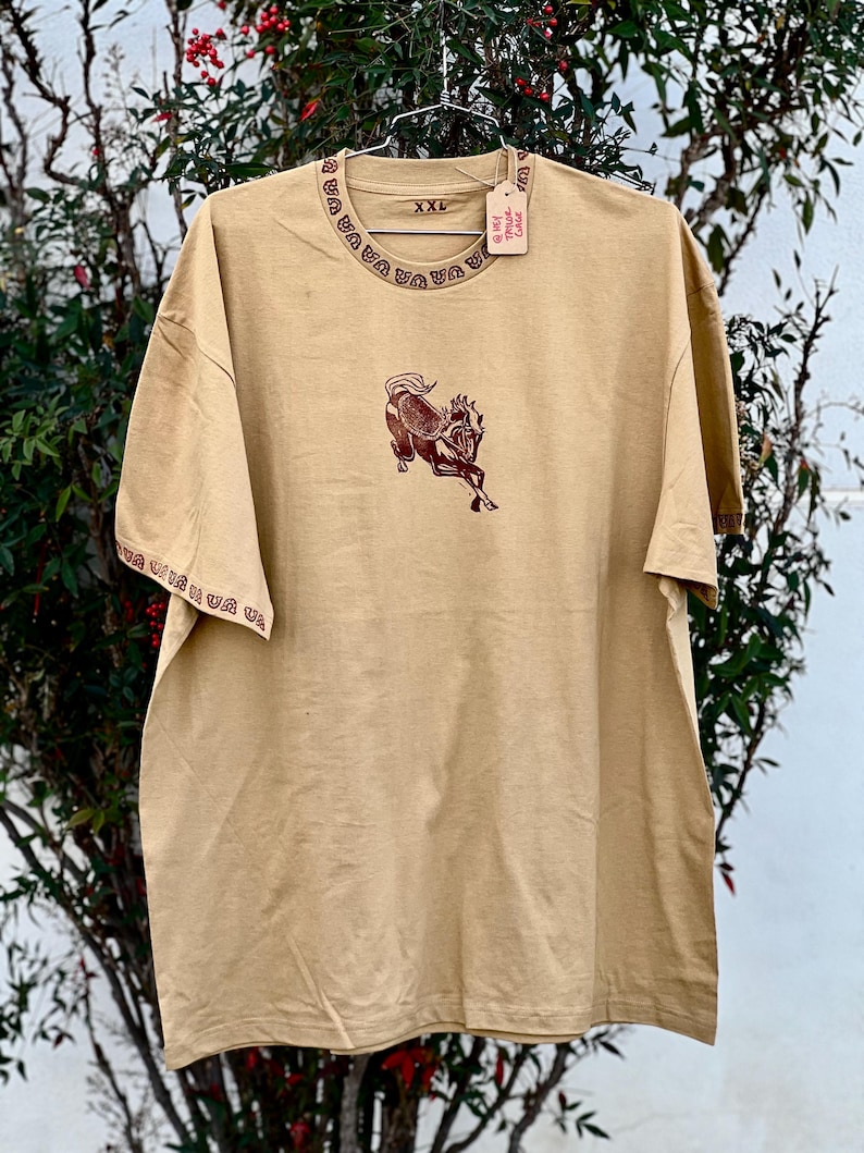 May include: A tan t-shirt with a brown horse graphic and a red trim around the neck and sleeves. The shirt has a tag that says "@Get True Cozy" and "XXL" printed on it.