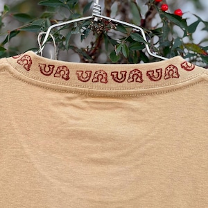 May include: A tan t-shirt with a red embroidered design around the neckline. The design is a repeating pattern of a horseshoe shape.