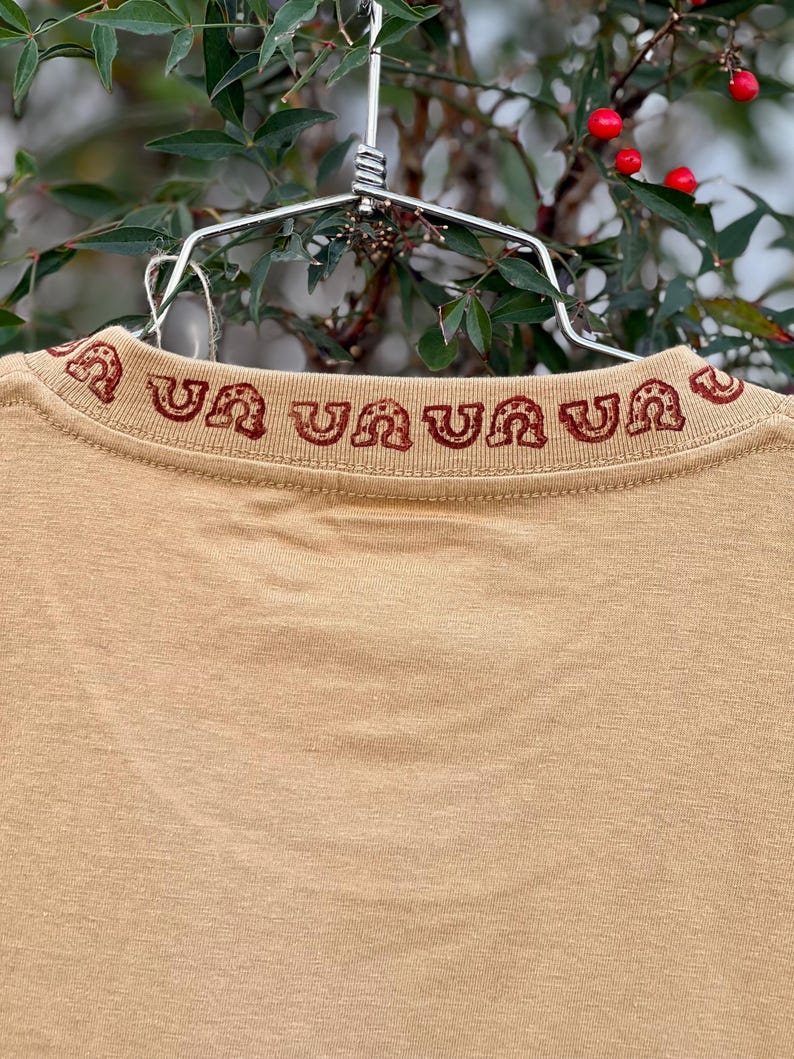 May include: A tan t-shirt with a red embroidered design around the neckline. The design is a repeating pattern of a horseshoe shape.