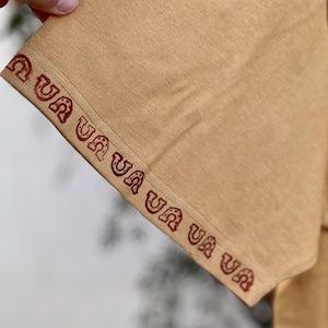 May include: Close-up of the tan fabric sleeve of a shirt with a red embroidered pattern that reads "Ua Ua Ua Ua Ua Ua".