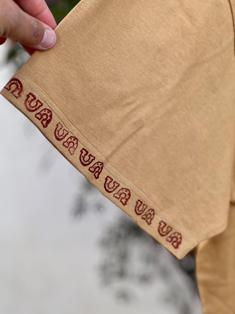 May include: Close-up of the tan fabric sleeve of a shirt with a red embroidered pattern that reads "Ua Ua Ua Ua Ua Ua".