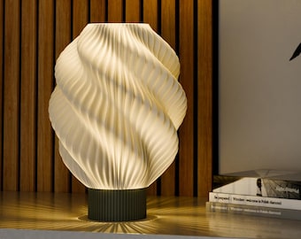 Könnte beinhalten: Eine weiße, spiralförmige Tischlampe mit grünem Fuß steht auf einer Holzoberfläche. Die Lampe ist eingeschaltet und strahlt ein warmes Licht aus. Hinter der Lampe liegen Bücher auf der Oberfläche.