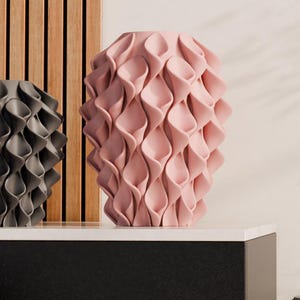 Op de afbeelding: Een verzameling decoratieve vazen in verschillende kleuren, waaronder roze, groen, grijs, zwart en koper. Elke vaas heeft een uniek, getextureerd ontwerp met een herhalend bloembladachtig patroon. De vazen staan op een wit oppervlak en een houten achtergrond.