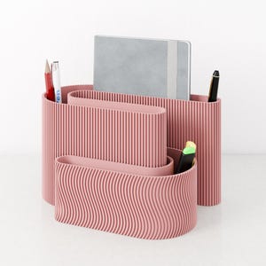 Könnte beinhalten: Ein rosa Schreibtisch-Organizer mit wellenförmigem und geripptem Design. Der Organizer enthält ein graues Notizbuch, Stifte und Bleistifte. Der Schreibtisch-Organizer besteht aus einem festen Material und ist für Büromaterialien konzipiert.