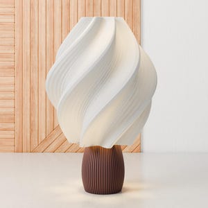 Op de afbeelding: Een witte tafellamp met een unieke, draaiende, sculpturale kap en een bruine, geribbelde voet.