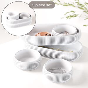 Könnte beinhalten: Ein 5-teiliges weißes Organizer-Set. Das Set enthält zwei lange, ovale Tabletts und drei runde Schalen. Die Tabletts und Schalen haben eine gerippte Textur. Das Set ist für kleine Gegenstände wie Schmuck und Bürobedarf konzipiert.