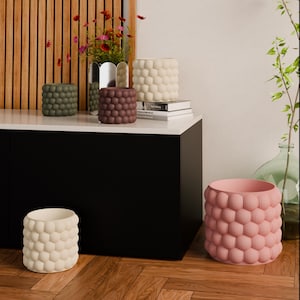 Può includere: Una collezione di vasi decorativi in vari colori, tra cui crema, rosa, verde oliva e bordeaux. I vasi hanno un design strutturato a bolle. Alcuni sono su una superficie bianca, altri sul pavimento.