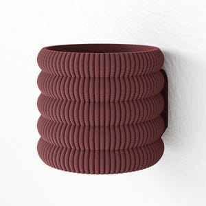 Könnte beinhalten: Ein maroonfarbener, gerippter Wandtopf. Der zylindrische Pflanztopf hat ein gestapeltes Design mit abgerundeten Rippen. Er ist auf einer dunkelmaroonfarbenen rechteckigen Basis an einer weißen Wand befestigt. Der Topf ist für den Innen- oder Außenbereich konzipiert.