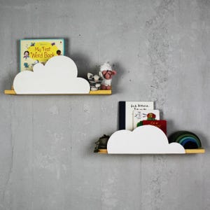 Pode incluir: Duas prateleiras brancas em forma de nuvem montadas em uma parede cinza. A prateleira superior tem um livro infantil intitulado "My First Word Book" e um animal de pelúcia. A prateleira inferior tem três livros e um brinquedo de empilhar arco-íris.