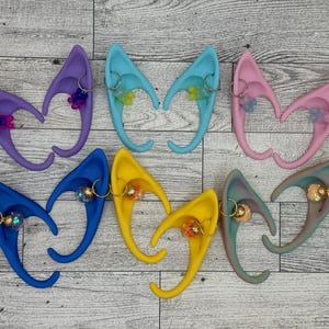 May include: Six pairs of elf ears in various colors, including purple, blue, yellow, and pink. Each pair has a small charm attached to the ear.