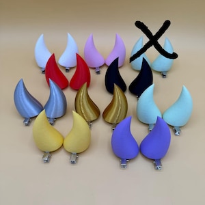 May include: A set of 12 pairs of devil horn hair clips in various colours, including red, white, pink, black, silver, gold, yellow, and purple. The horns are made of a smooth, plastic-like material and have a clip on the back for easy attachment to hair.
