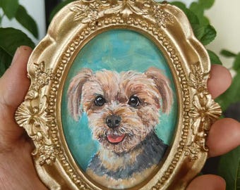 May include: A small, oval-shaped painting of a dog in a gold-colored ornate frame. The dog has brown and black fur, with a pink tongue. The background is a light blue color. The frame has a detailed, decorative design.