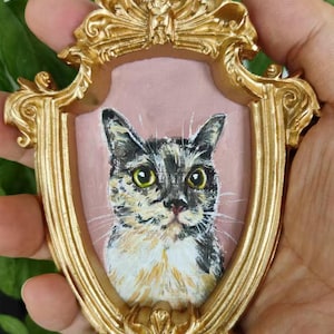 Puede incluir: Un pequeño marco dorado adornado con un retrato de gato. El gato tiene pelaje negro, blanco y marrón y ojos amarillos. El fondo del retrato es rosa. El marco tiene un diseño decorativo detallado.