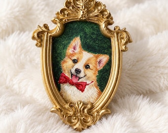 May include: A decorative wall plaque featuring a detailed embroidery of a tan and white Corgi dog wearing a bright red bow tie. The portrait is encased in an ornate, metallic gold-colored frame with intricate scrollwork and a dark green background.