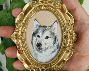 May include: A small, oval-shaped painting of a dog in a gold ornate frame. The dog has black and gray fur with a white face and chest. The frame is a detailed, antique-style design with floral accents. The background of the painting is a light tan color.