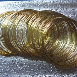 May include: A collection of coiled, golden-colored wire, arranged in a circular pattern. The wire appears to be made of a shiny metal, reflecting light and creating highlights. The coils are stacked, creating a layered effect. The background is a textured, silver surface.