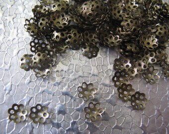 May include: A pile of small, gold-colored metal flower-shaped embellishments with a decorative openwork design. The embellishments are scattered on a textured silver surface.