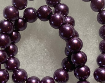 May include: A strand of dark purple, round beads. The beads have a glossy finish and are strung together on a thin, clear string. The beads are evenly sized and spaced, creating a consistent pattern.
