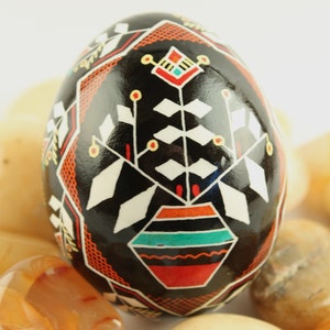 May include: A black egg with a white, red and yellow geometric design. The egg is decorated with a stylised flower and a pot with a turquoise and red design.