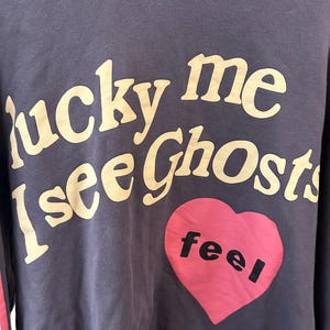 May include: A navy blue hoodie with the text "lucky me I see Ghosts" in a cream-colored font. A pink heart with the word "feel" in black is on the front. The hoodie has a hood and a red and white stripe on the sleeve.