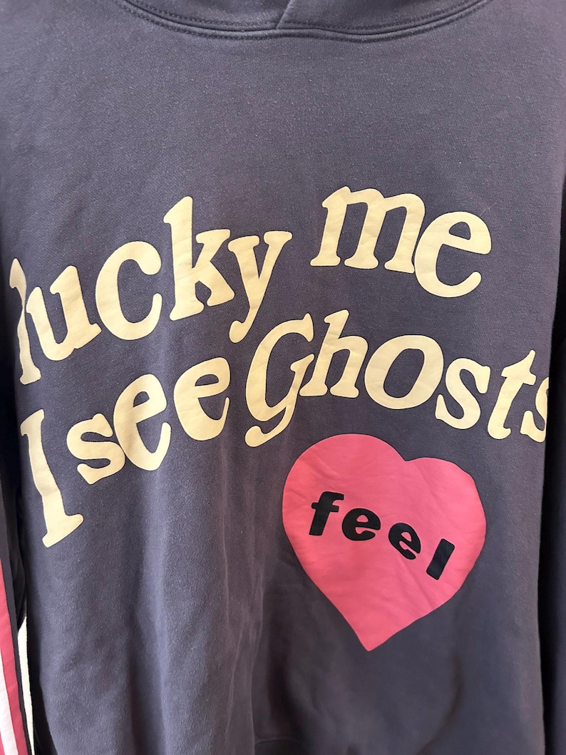 May include: A navy blue hoodie with the text "lucky me I see Ghosts" in a cream-colored font. A pink heart with the word "feel" in black is on the front. The hoodie has a hood and a red and white stripe on the sleeve.
