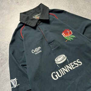 May include: Dark navy rugby shirt with a black collar and red trim. Features a red rose emblem, the Guinness logo, and the text "Cotton Traders". The shirt has short sleeves and a button-down collar.