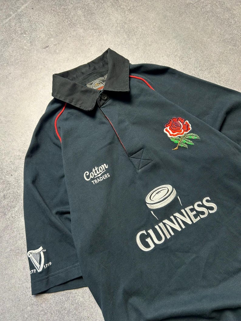 May include: Dark navy rugby shirt with a black collar and red trim. Features a red rose emblem, the Guinness logo, and the text "Cotton Traders". The shirt has short sleeves and a button-down collar.