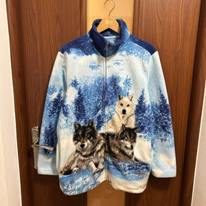 May include: A full-zip fleece jacket featuring a winter landscape with three wolves. The jacket has a blue and white color scheme with a high collar and a zipper closure. The wolves are depicted in shades of gray and white.