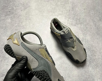 May include: A pair of gray Puma sneakers with black soles. The shoes have a textured design and a strap closure. The Puma logo is visible on the side and tongue of the shoe. The shoes are shown on a gray surface.