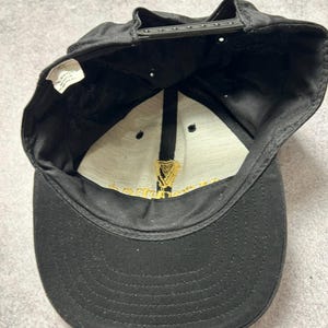 Pu&ograve; includere: Cappellino da baseball nero con visiera curva e chiusura a scatto regolabile. L'interno presenta un pannello bianco con testo dorato e un'emblema dorato dell'arpa. L'esterno del cappellino &egrave; nero.