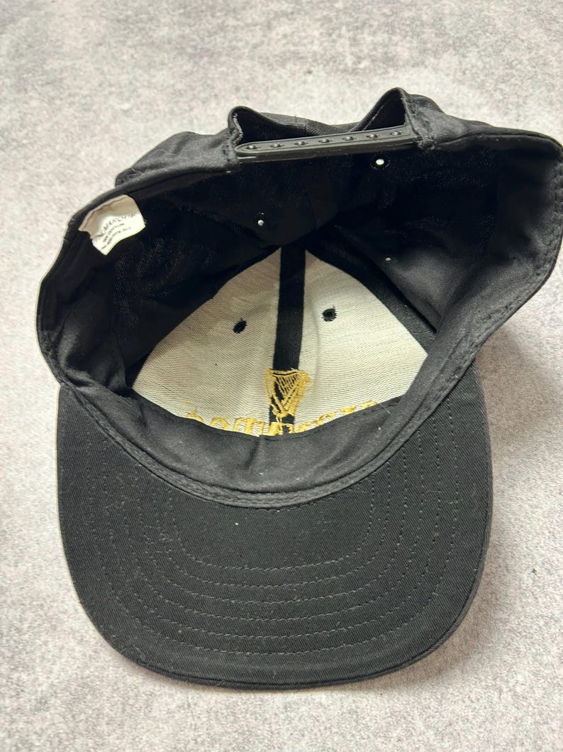 Pu&ograve; includere: Cappellino da baseball nero con visiera curva e chiusura a scatto regolabile. L'interno presenta un pannello bianco con testo dorato e un'emblema dorato dell'arpa. L'esterno del cappellino &egrave; nero.
