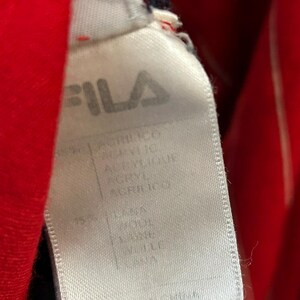 Puede incluir: Una etiqueta de ropa de tela blanca con el logotipo de la marca FILA en texto negro en la parte superior. La etiqueta enumera la composici&oacute;n del material, incluyendo acr&iacute;lico y 15% lana, con el texto Made in China impreso en el borde inferior.