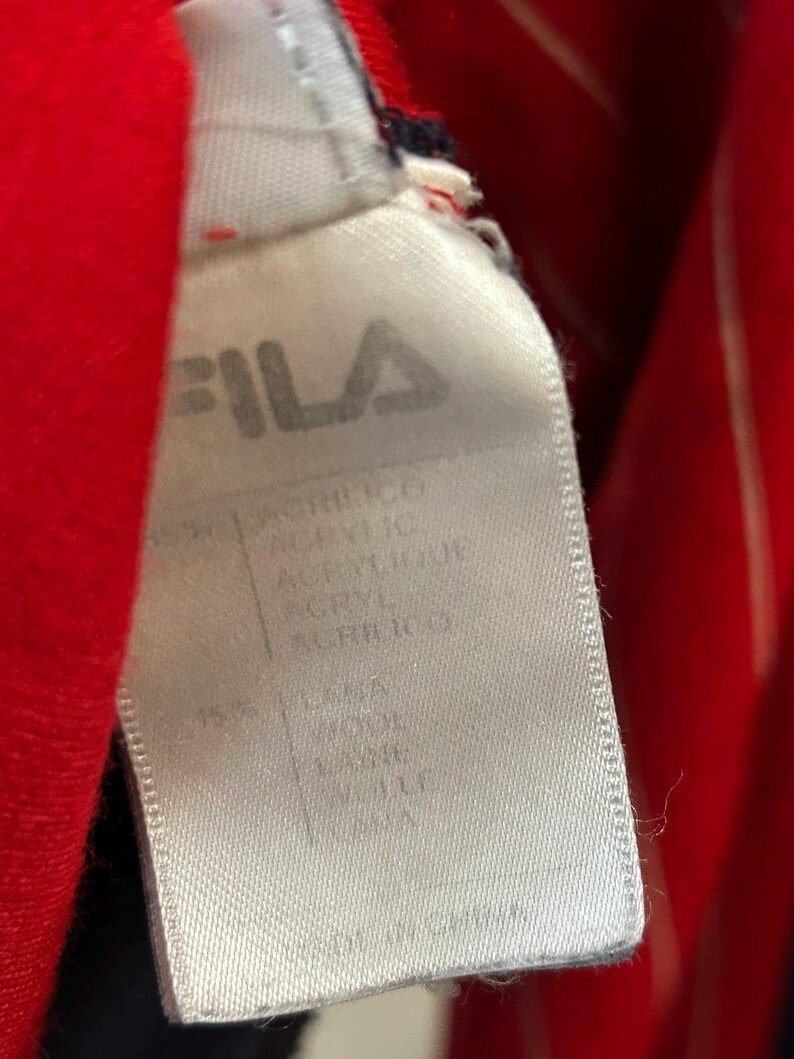 Puede incluir: Una etiqueta de ropa de tela blanca con el logotipo de la marca FILA en texto negro en la parte superior. La etiqueta enumera la composici&oacute;n del material, incluyendo acr&iacute;lico y 15% lana, con el texto Made in China impreso en el borde inferior.