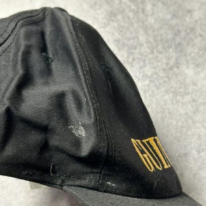 Pu&ograve; includere: Cappellino da baseball nero con visiera curva. Il cappellino presenta un testo dorato ricamato sul lato. Il cappellino &egrave; realizzato in tessuto strutturato e presenta segni di usura.
