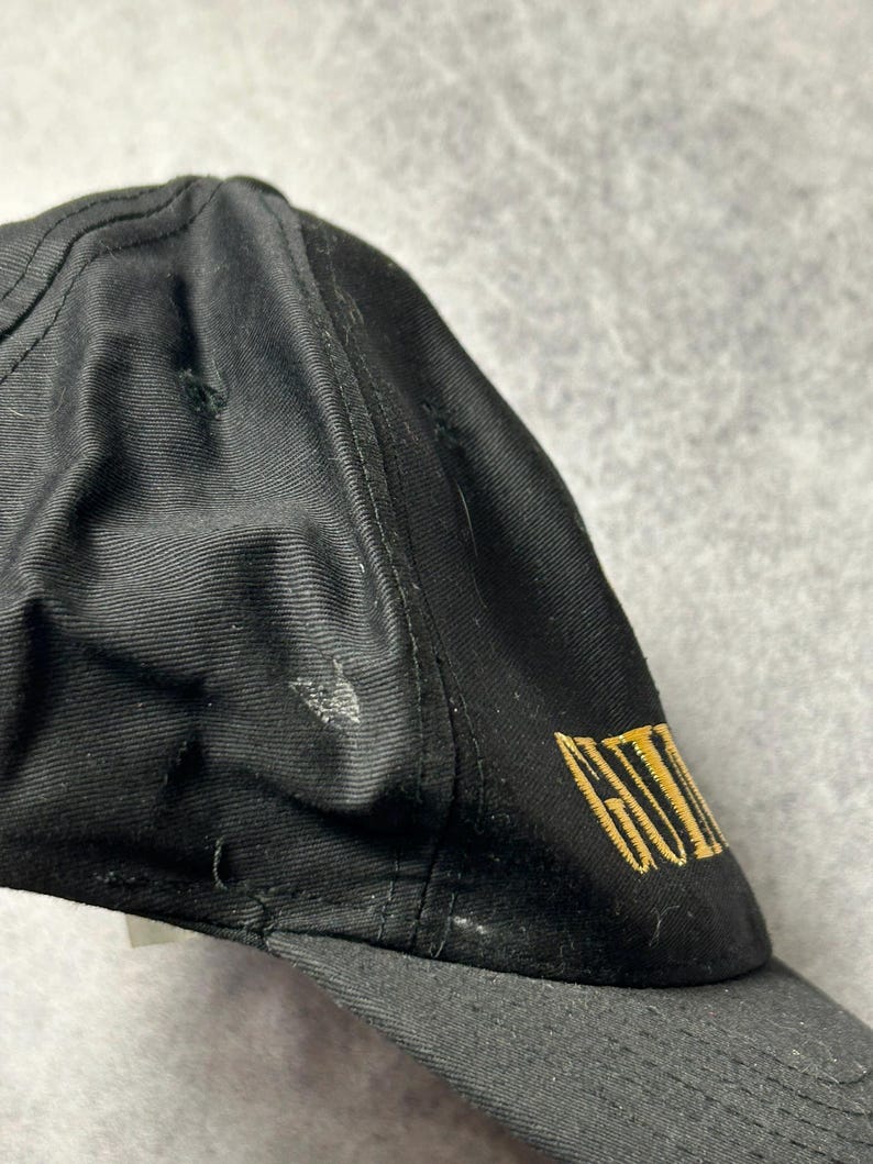 Pu&ograve; includere: Cappellino da baseball nero con visiera curva. Il cappellino presenta un testo dorato ricamato sul lato. Il cappellino &egrave; realizzato in tessuto strutturato e presenta segni di usura.