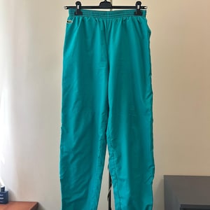 Op de afbeelding: Teal kleurige trainingsbroek met een elastische tailleband en een klein geborduurd krokodillenlogo op de linkerheup. De broek heeft een relaxte pasvorm met elastische boorden bij de enkels en is gemaakt van een gladde, lichtgewicht stof.