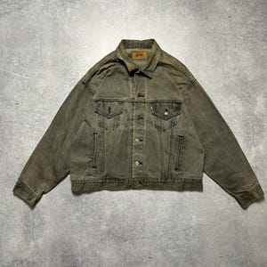 May include: A vintage green denim jacket with a button closure and two front pockets. The jacket has a faded look and a worn-in feel.