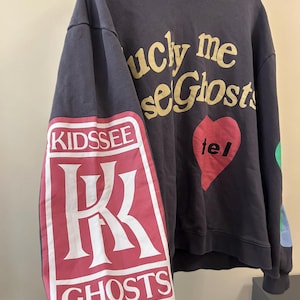 May include: Dark gray hoodie with the text "lucky me see Ghosts" in gold lettering. The left sleeve features a red and white graphic with the words "KIDS SEE GHOSTS". A red heart with the word "tel" is on the front.