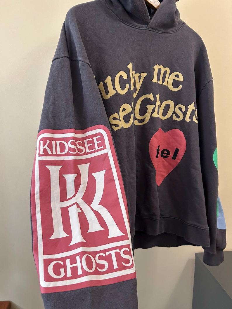 May include: Dark gray hoodie with the text "lucky me see Ghosts" in gold lettering. The left sleeve features a red and white graphic with the words "KIDS SEE GHOSTS". A red heart with the word "tel" is on the front.